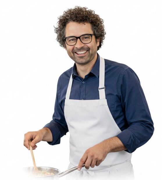 Ein freundlich lächelnder Mann mit Brille und dunklen Locken trägt eine weiße Kochschürze über einem blauen Hemd. Er steht vor einem weißen Hintergrund und rührt mit einem Holzlöffel in einer Pfanne.